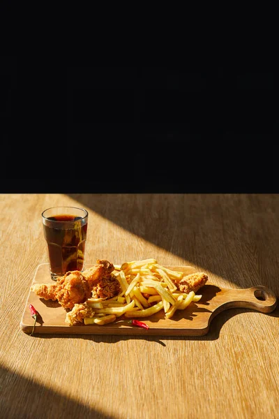 Pollo fritto speziato, patatine fritte a bordo con soda in vetro su tavolo di legno alla luce del sole isolato su nero — Foto stock
