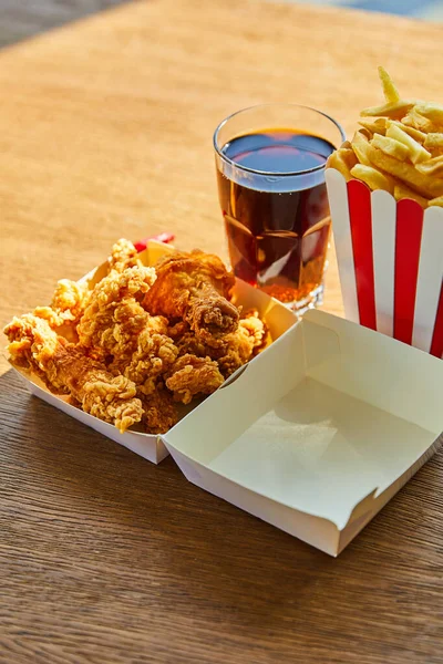 Deep fried chicken, french fries and soda in glass on wooden table in sunlight — Stock Photo