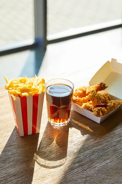 Spicy deep fried chicken, french fries and soda in glass on wooden table in sunlight near window — Stock Photo