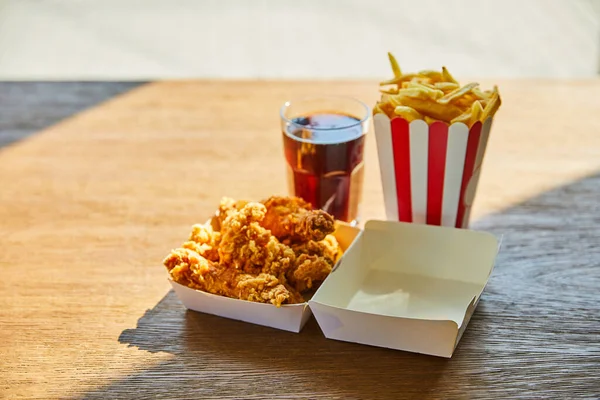 Fuoco selettivo di pollo fritto in profondità, patatine fritte e soda in vetro su tavolo di legno alla luce del sole vicino alla finestra — Foto stock