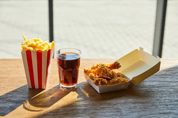 Pollo frito picante, papas fritas y refrescos en vidrio sobre mesa de madera a la luz del sol cerca de la ventana - foto de stock