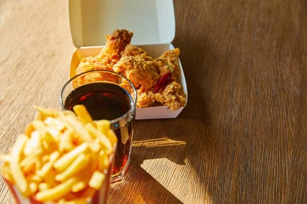 Fuoco selettivo di pollo fritto speziato, patatine fritte e soda in vetro sul tavolo di legno alla luce del sole — Foto stock