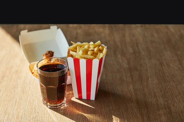 Deep fried chicken, french fries and soda in glass on wooden table in sunlight isolated on black — Stock Photo