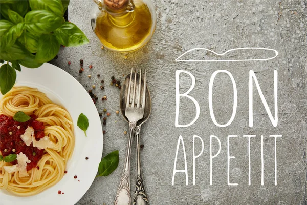 Top view of delicious spaghetti with tomato sauce on plate near basil leaves, oil and cutlery on grey textured surface with bon appetit illustration — Stock Photo