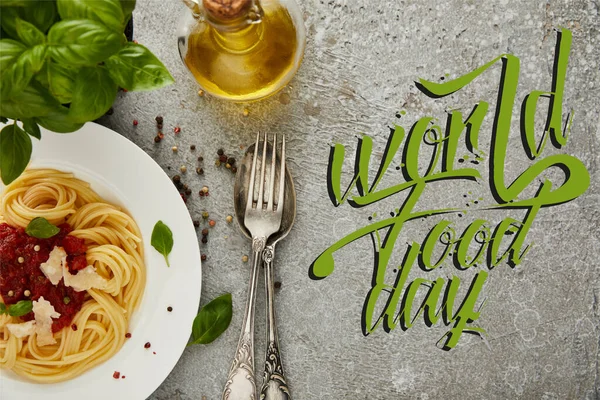 Vue du dessus de délicieux spaghettis à la sauce tomate sur une assiette près des feuilles de basilic, de l'huile et des couverts sur une surface grise texturée avec illustration de la journée mondiale de l'alimentation — Photo de stock