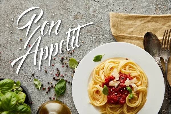 Top view of delicious spaghetti with tomato sauce on plate near basil leaves and cutlery on grey textured surface with bon appetit illustration — Stock Photo