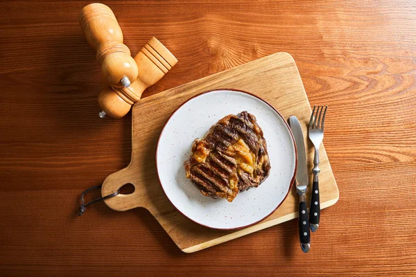 Blick von oben auf frisch gegrilltes Steak auf Teller auf Schneidebrett mit Besteck und Salz- und Pfeffermühlen auf Holztisch — Stockfoto