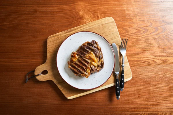 Draufsicht auf frisch gegrilltes Steak auf Teller auf Schneidebrett mit Besteck auf Holztisch — Stockfoto