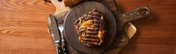 Top view of tasty grilled steak served on wooden board near cutlery, panoramic shot — Stock Photo