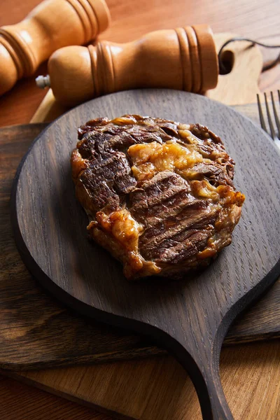 Selective focus of tasty grilled steak served on wooden board near salt and pepper mills — Stock Photo