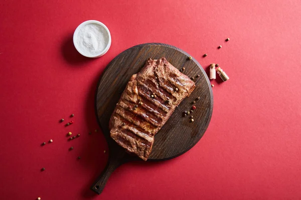 Top view of tasty grilled steak served on wooden board on red background with salt and pepper — Stock Photo