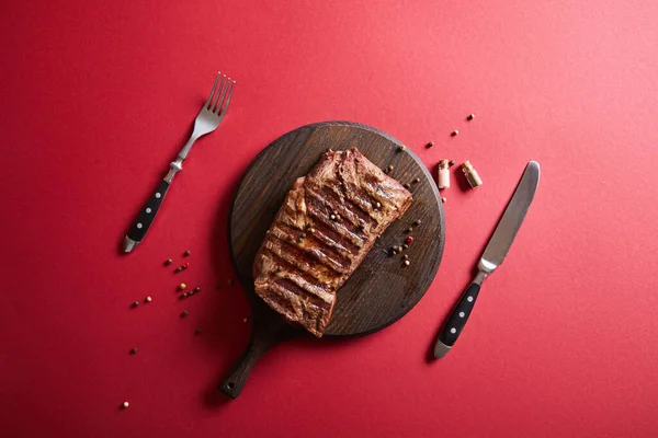 Vue de dessus de steak grillé savoureux servi sur une planche de bois sur fond rouge avec poivre et couverts — Photo de stock