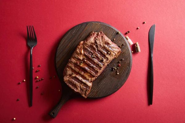 Vista superior de saboroso bife grelhado servido em tábua de madeira em fundo vermelho com pimenta e talheres — Fotografia de Stock