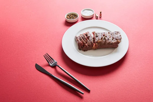 Tasty grilled steak served on plate with cutlery and salt and pepper in bowls on red background — Stock Photo