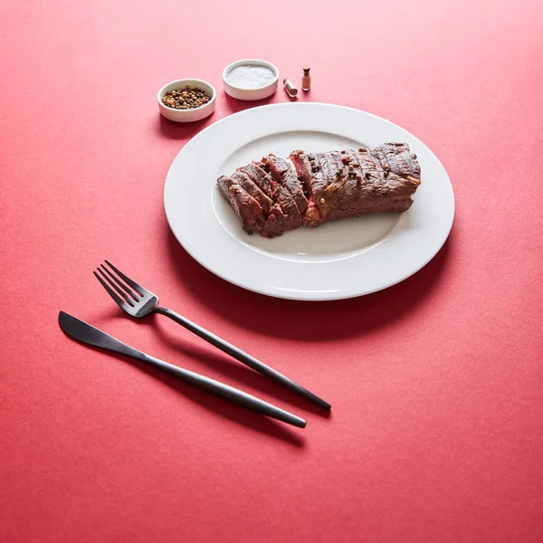 Leckeres Steak vom Grill serviert auf Teller mit Besteck und Salz und Pfeffer in Schüsseln auf rotem Hintergrund — Stockfoto