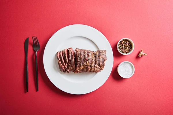 Top view of tasty grilled steak served on plate with cutlery and salt and pepper in bowls on red background — Stock Photo