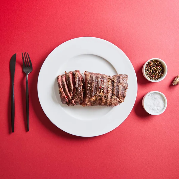 Top view of tasty grilled steak served on plate with cutlery and salt and pepper in bowls on red background — Stock Photo