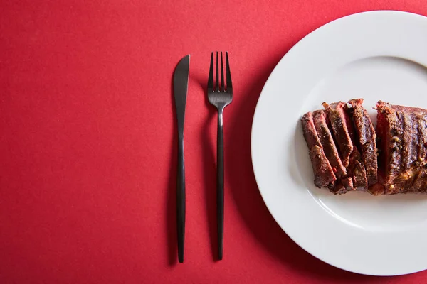 Blick von oben auf leckeres Steak vom Grill serviert auf Teller mit Besteck auf rotem Hintergrund — Stockfoto