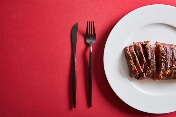 Вид сверху на вкусный стейк на гриле подается на тарелке со столовыми приборами на красном фоне — стоковое фото