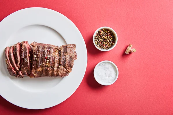 Top view of tasty grilled steak served on plate with salt and pepper in bowls on red background — Stock Photo