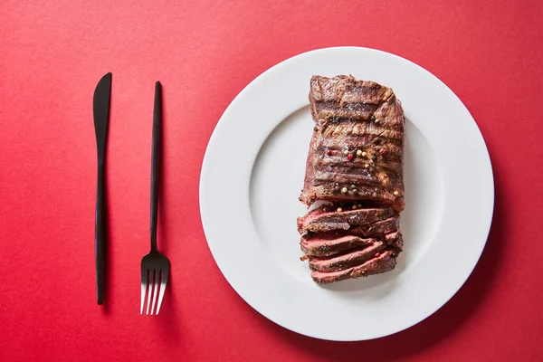 Blick von oben auf leckeres Steak vom Grill serviert auf Teller mit Besteck auf rotem Hintergrund — Stockfoto