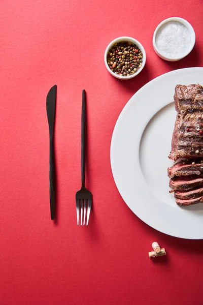 Top view of tasty grilled steak served on plate with cutlery and salt and pepper in bowls on red background — Stock Photo