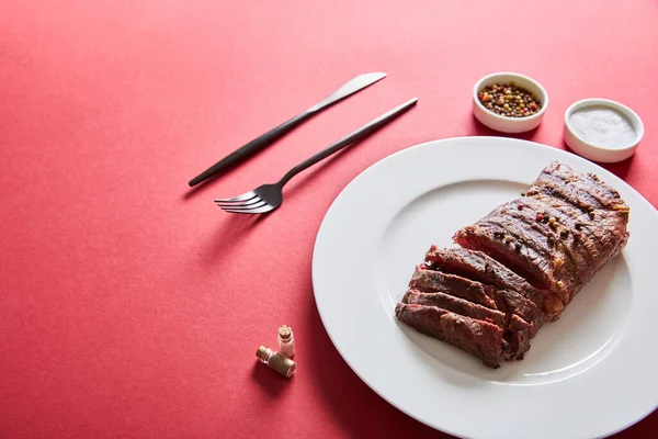 Tasty grilled steak served on plate with cutlery and salt and pepper in bowls on red background — Stock Photo