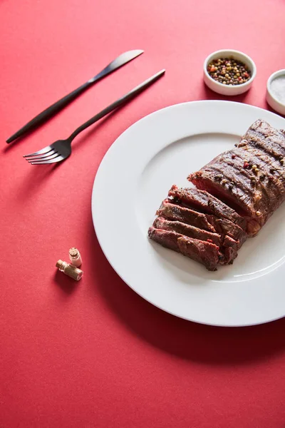 Saboroso bife grelhado servido em prato com talheres e sal e pimenta em tigelas em fundo vermelho — Fotografia de Stock