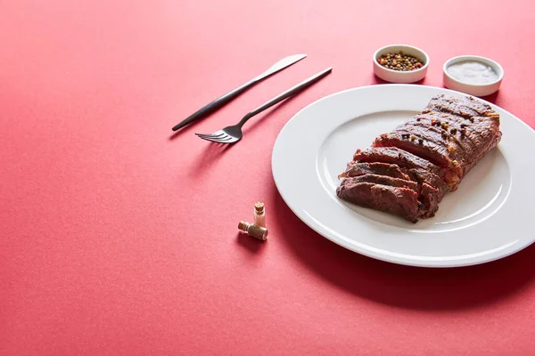 Savoureux steak grillé servi dans une assiette avec couverts et sel et poivre dans des bols sur fond rouge — Photo de stock