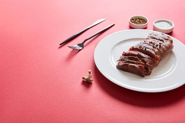 Saboroso bife grelhado servido em prato com talheres e sal e pimenta em tigelas em fundo vermelho — Fotografia de Stock