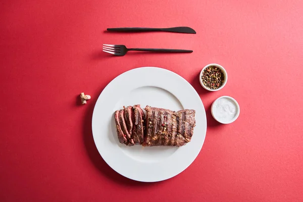 Top view of tasty grilled steak served on plate with cutlery and salt and pepper in bowls on red background — Stock Photo