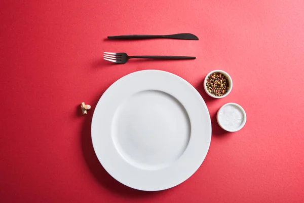 Вид сверху на пустую круглую тарелку со столовыми приборами и солью и перцем в мисках на красном фоне — стоковое фото