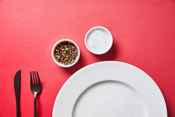 Вид зверху порожня кругла тарілка з столовими приборами, сіллю та перцем у мисках на червоному тлі — Stock Photo