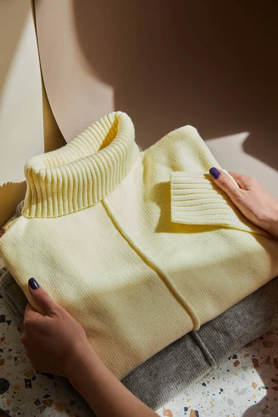 Vista recortada de la mujer sosteniendo suéteres suaves de punto amarillo y gris en la superficie de piedra y papel a la luz del sol - foto de stock