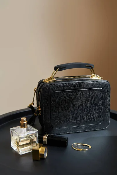 Leather handbag near golden earrings, perfume and lipstick on black table on beige background — Stock Photo