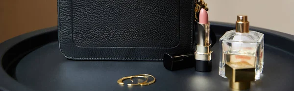 Leather handbag near golden earrings, perfume and lipstick on black table, panoramic shot — Stock Photo