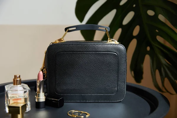 Bolsa de couro perto de brincos dourados, perfume e batom na mesa preta perto de folha tropical — Fotografia de Stock