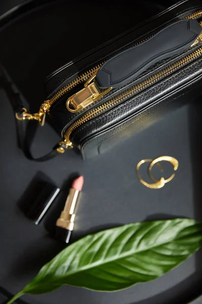 Top view of leather handbag near golden earrings and lipstick on black table with green leaf — Stock Photo
