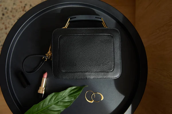 Vue du dessus du sac à main en cuir près de boucles d'oreilles dorées et rouge à lèvres sur table noire avec feuille tropicale — Photo de stock