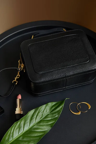 Leather handbag near golden earrings and pink lipstick on black table with tropical leaf — Stock Photo