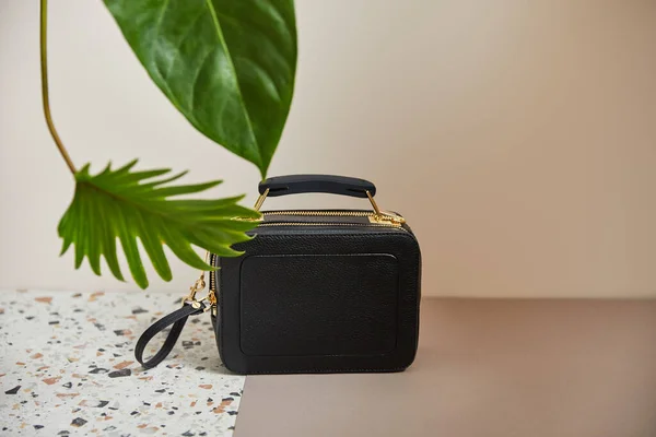 Leather black handbag with golden zippers on marble surface on beige background with tropical green leaves — Stock Photo