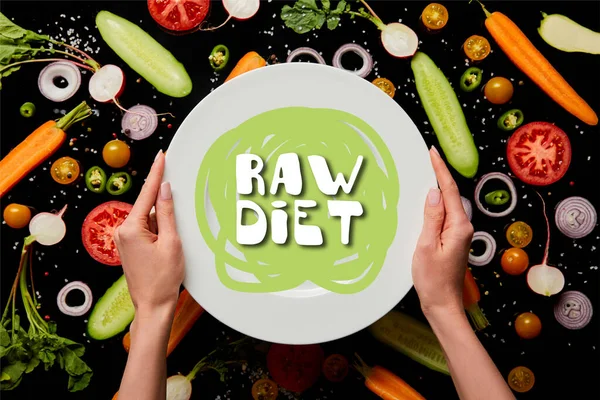 Cropped view of woman holding round plate with raw diet illustration on vegetable pattern background isolated on black — Stock Photo