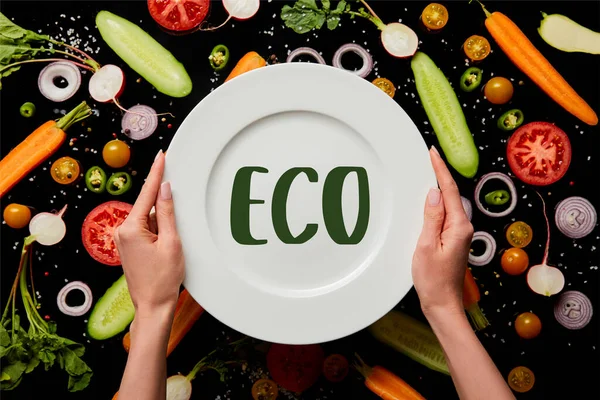 Cropped view of woman holding round plate with eco illustration on vegetable pattern background isolated on black — Stock Photo