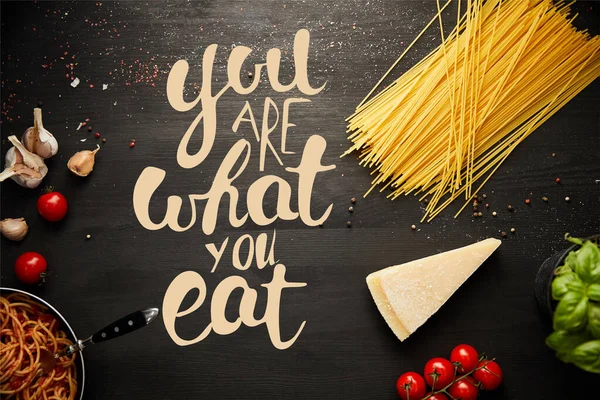 Top view of tasty bolognese pasta in frying pan near ingredients on black wooden background with you are what you eat illustration — Stock Photo