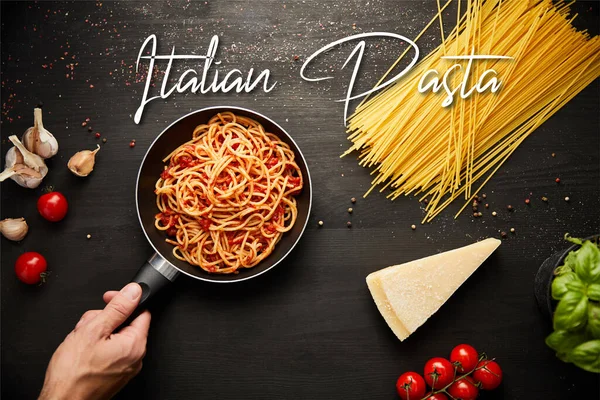 Cropped view of man holding frying pan with tasty bolognese pasta near ingredients on black wooden background, italian pasta illustration — Stock Photo