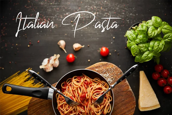 Top view of tasty bolognese pasta in frying pan near ingredients and cutlery on black wooden background, italian pasta illustration — Stock Photo