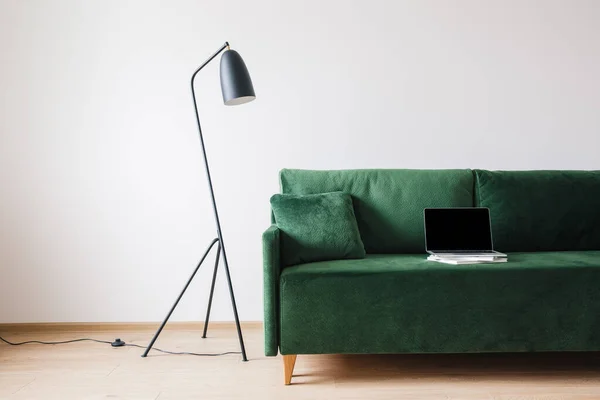Sofá verde com travesseiro e laptop com tela em branco em livros perto de metal lâmpada de assoalho moderna — Fotografia de Stock