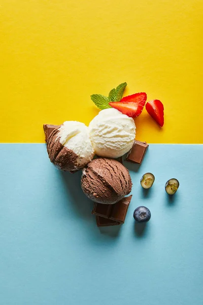 Draufsicht auf leckeres braunes und weißes Eis mit Beeren, Schokolade und Minze auf gelbem und blauem Hintergrund — Stockfoto