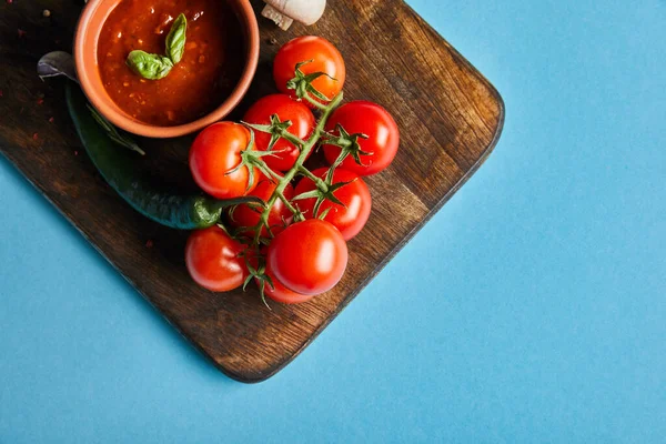 Vista superior de delicioso molho de tomate na tigela em tábua de madeira com legumes maduros frescos no fundo azul — Fotografia de Stock