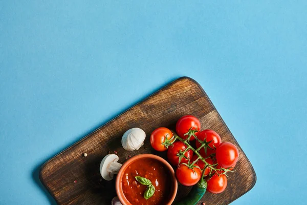 Вид на вкусный томатный соус в миске на деревянной доске со свежими спелыми овощами на синем фоне — стоковое фото